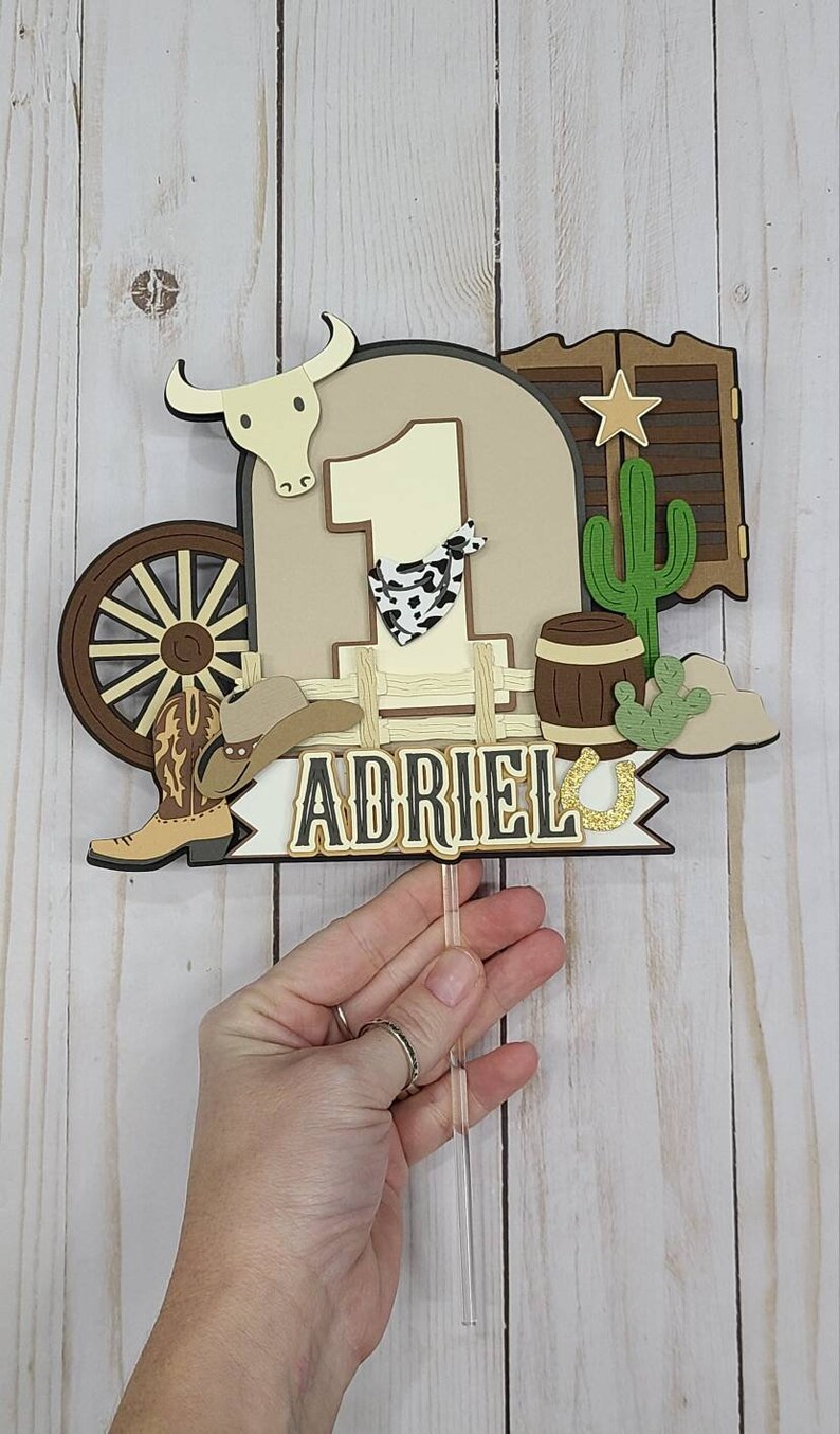 May include: A brown, white, and gold cowboy-themed cake topper with the number 1, a cowboy boot, a cowboy hat, a wagon wheel, a cactus, a barrel, and a horseshoe. The name "ADRIEL" is written on the topper.