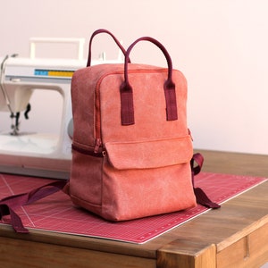 May include: A coral-colored canvas backpack with burgundy straps and a zippered front pocket. The backpack is sitting on a red cutting mat on a wooden table.