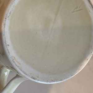May include: The bottom of a cream-colored ceramic mug with a small, handwritten mark on the bottom.