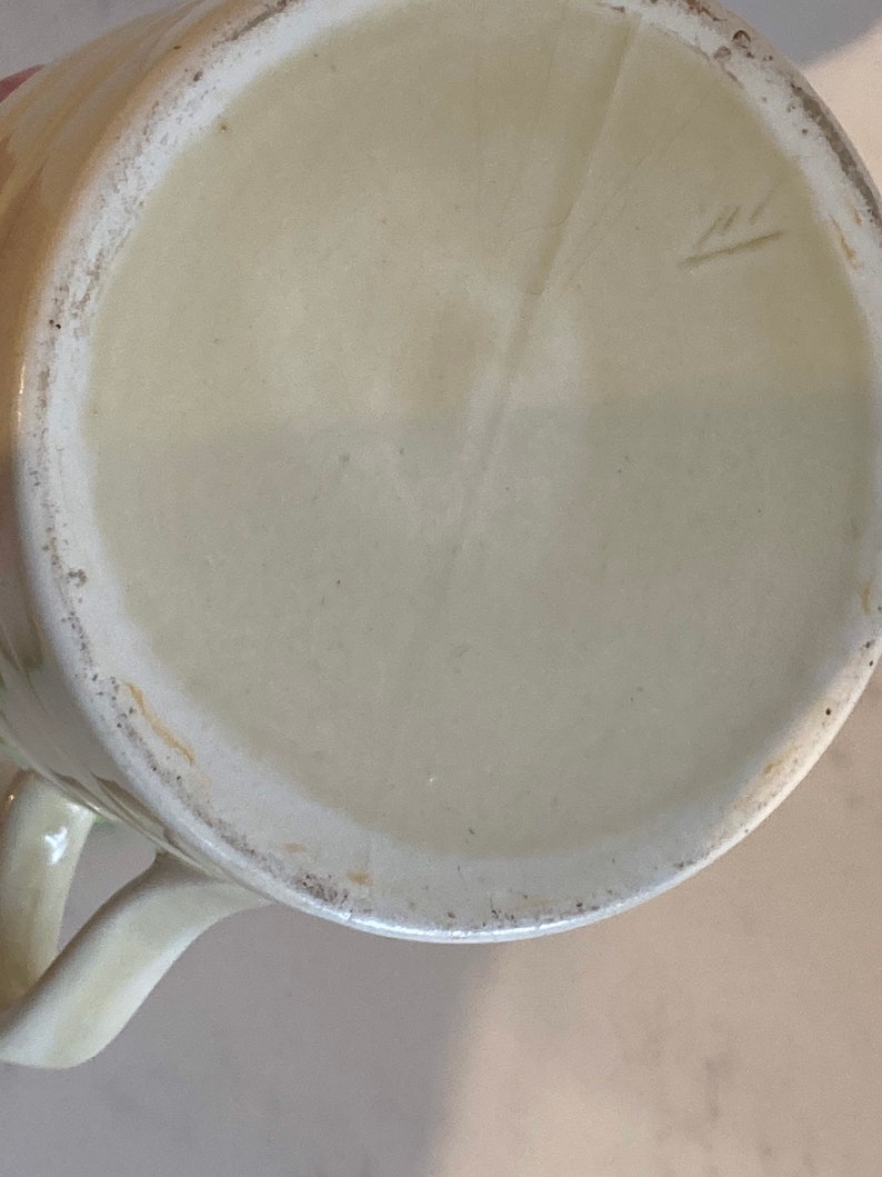 May include: The bottom of a cream-colored ceramic mug with a small, handwritten mark on the bottom.
