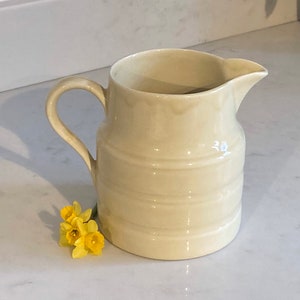 May include: A cream-colored ceramic pitcher with a ribbed design and a handle. The pitcher is sitting on a white countertop.