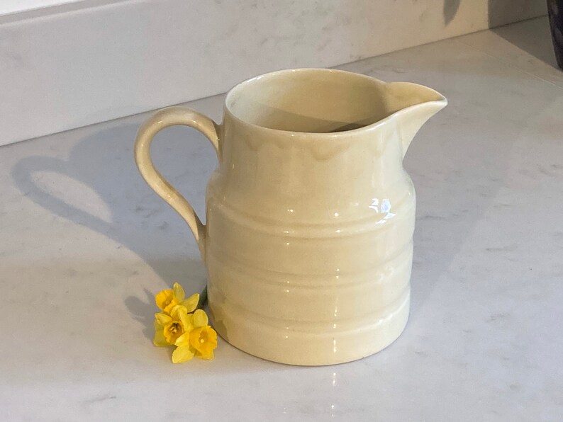 May include: A cream-colored ceramic pitcher with a ribbed design and a handle. The pitcher is sitting on a white countertop.