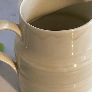 May include: A white ceramic pitcher with a rounded body and a handle. The pitcher has a smooth, glossy finish and is filled with a light brown liquid.