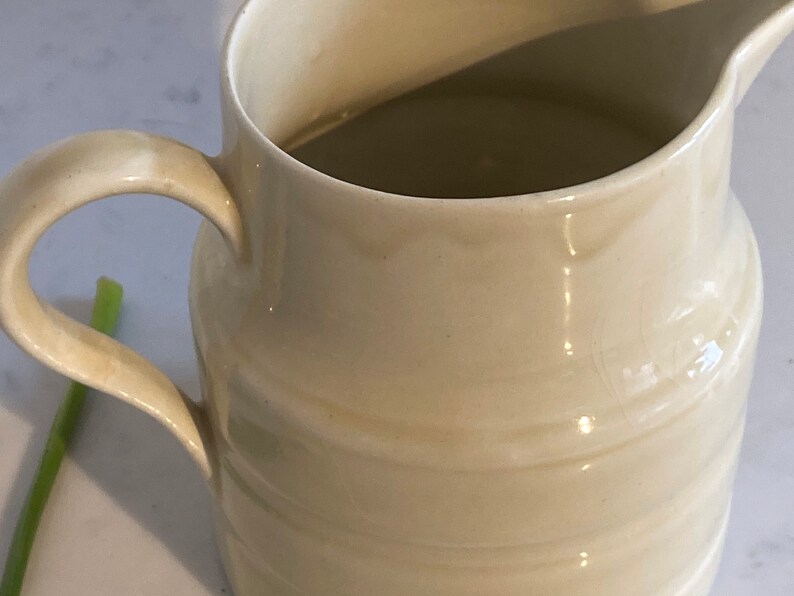 May include: A white ceramic pitcher with a rounded body and a handle. The pitcher has a smooth, glossy finish and is filled with a light brown liquid.