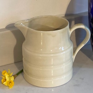 May include: A white ceramic pitcher with a rounded body and a handle. The pitcher has a smooth, glossy finish and a slightly textured design.