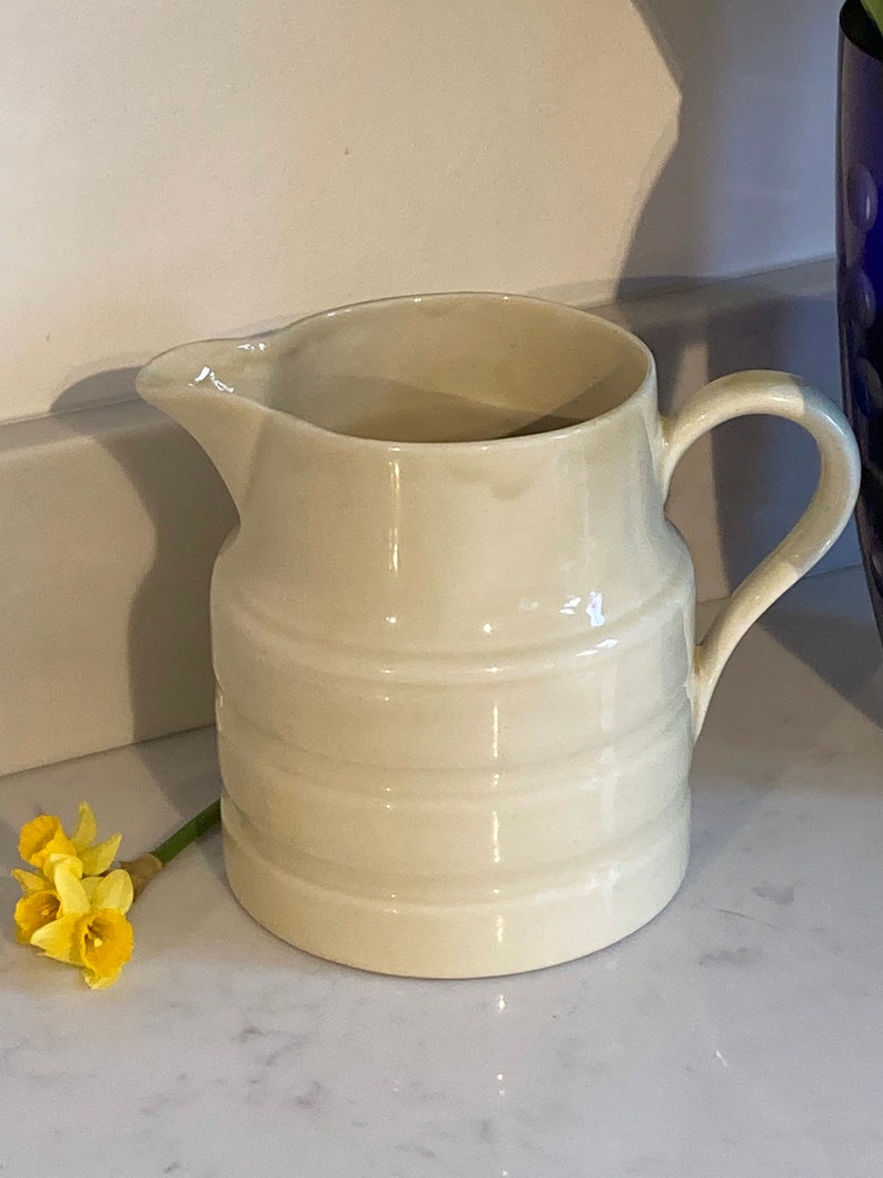May include: A white ceramic pitcher with a rounded body and a handle. The pitcher has a smooth, glossy finish and a slightly textured design.