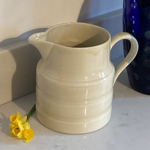 May include: A white ceramic pitcher with a ribbed design. The pitcher has a handle and a spout. It is sitting on a white countertop.