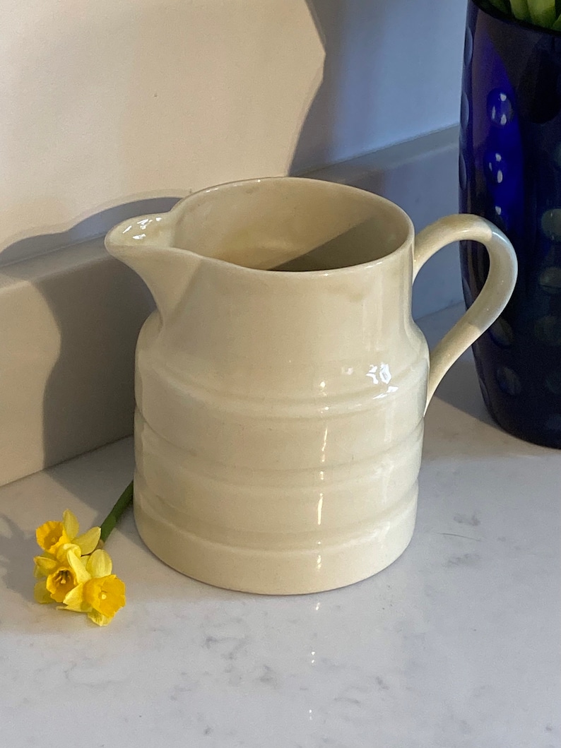 May include: A white ceramic pitcher with a ribbed design. The pitcher has a handle and a spout. It is sitting on a white countertop.