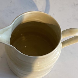 May include: A cream-colored ceramic pitcher with a rounded body and a slightly flared lip. The pitcher has a handle and a textured, ribbed design.