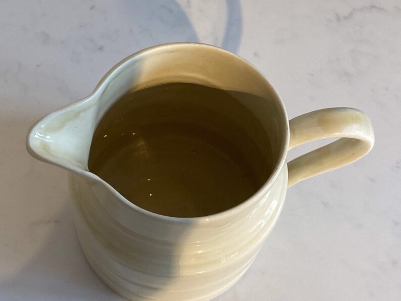 May include: A cream-colored ceramic pitcher with a rounded body and a slightly flared lip. The pitcher has a handle and a textured, ribbed design.