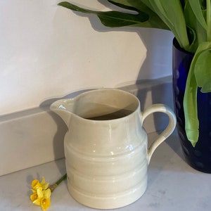 May include: A white ceramic pitcher with a ribbed design and a handle. The pitcher is sitting on a white countertop.