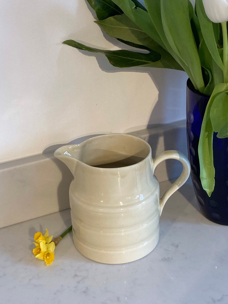 May include: A white ceramic pitcher with a ribbed design and a handle. The pitcher is sitting on a white countertop.