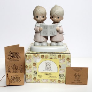May include: A Precious Moments figurine depicting two children singing from a music sheet. The figurine is on a decorative base, atop a box with angel illustrations. Additional tags and packaging are included, with the text "Peace On Earth".