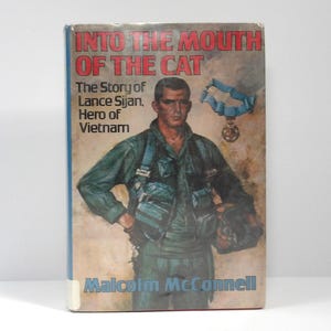 May include: A hardcover book titled Into the Mouth of the Cat: The Story of Lance Sijan, Hero of Vietnam by Malcolm McConnell. The cover features a painted illustration of a pilot in a green flight suit holding a helmet with a medal above.