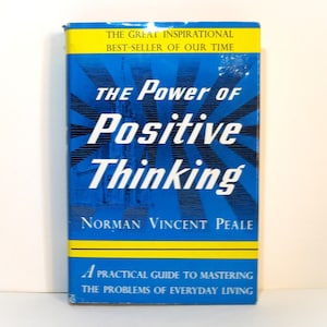 May include: A book titled The Power of Positive Thinking by Norman Vincent Peale. The cover is blue with yellow horizontal bands at the top and bottom. It features white text and a sunburst graphic design behind the title and author name.