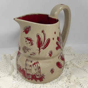May include: A ceramic jug with a beige exterior and a vibrant red interior. The outer surface features a hand-painted red design depicting a figure wearing a hat and striped clothing, surrounded by stylised floral and leaf motifs.