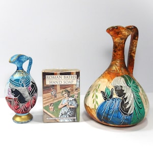 May include: Two ceramic pitchers with painted figures and a rectangular box of Roman Baths hand soap. The smaller pitcher is blue and red, while the larger one is brown, green, and blue. The soap box features a classical bath scene.