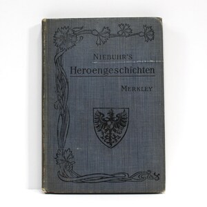 May include: A vintage book with a blue cover featuring the title "NIEBUHR'S Heroengeschichten" and the name "Merkley." The cover has decorative floral elements and a crest with an eagle. The book's edges show signs of wear.