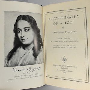 May include: An open hardcover book titled Autobiography of a Yogi by Paramahansa Yogananda. The left page features a black and white portrait of the author with a signature below. The right page displays the title, author, and publication details.