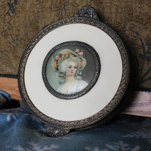 Puede incluir: Un marco de retrato redondo y antiguo con una pintura en miniatura de una mujer con sombrero. El marco tiene un centro color crema y un borde de metal ornamentado. La pintura presenta a una mujer con peluca empolvada y un sombrero adornado con rosas rosas.