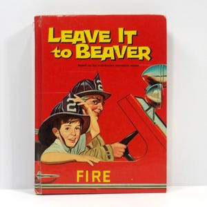 Puede incluir: Una portada de libro vintage de "Leave It to Beaver". La portada es roja con texto amarillo y presenta una ilustración de un niño y un hombre mayor en un camión de bomberos. La palabra "FIRE" está impresa en la parte inferior.