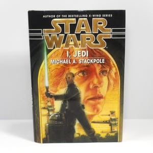 May include: A hardcover book titled "Star Wars I, Jedi" by Michael A. Stackpole. The cover features a man holding a green lightsaber, with a portrait of a person in the background. The Star Wars logo is at the top.