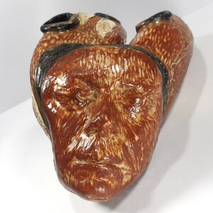 May include: A ceramic sculpture of a face with three openings. The sculpture is primarily brown with black and tan accents. The face has detailed features and a textured surface, suggesting a handcrafted piece of art.