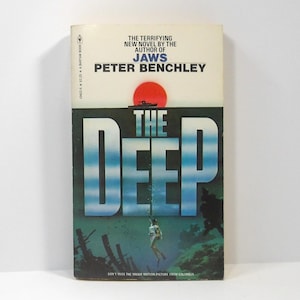 May include: A paperback book titled "The Deep" by Peter Benchley. The cover features a diver, the title in large blue letters, and a red sun. The text reads "The terrifying new novel by the author of Jaws".