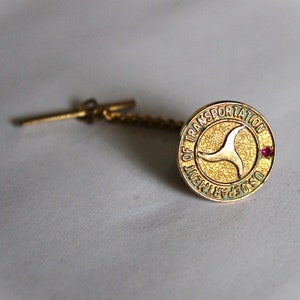 May include: Gold tie tack with a green and gold circular design featuring the text "U.S. Department of Transportation" and a gold stylized arrow. A small red stone is embedded in the design.