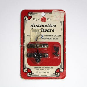 May include: A red and white card with the words "distinctive hardware" printed on it. The card features a small black metal latch with three black balls and two gold metal plates. The card also includes two small screws. The text on the card reads "ACL Pewter Catch Screws/Price $1.25".