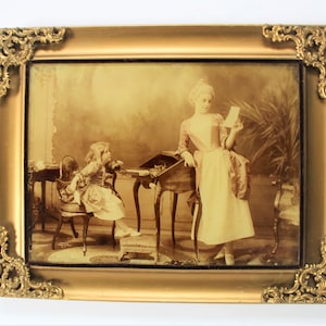 May include: A sepia-toned photograph in a gold frame shows two girls in a Victorian-era setting. The older girl is standing and holding a letter, while the younger girl is seated at a desk. The photograph is likely from the late 19th century.