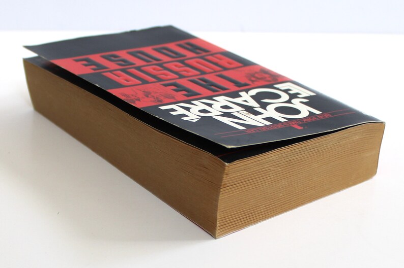 May include: A brown paperback book with the title "The Care of the Russian House" by John le Carre. The book is closed and the cover is slightly bent.