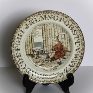 May include: A white ceramic plate with a brown and blue illustration of a man working on a wooden structure. The plate has a brown border with the letters of the alphabet in a circle around the edge. The text "CRUSOE AT WORK" is printed below the illustration.