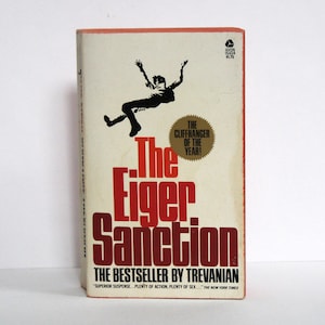 Puede incluir: Un libro de bolsillo rojo y blanco con el título "The Eiger Sanction" en letras rojas grandes y en negrita. La portada del libro presenta una silueta negra de una persona cayendo de un acantilado. El texto "The Bestseller by Trevanian" está impreso debajo del título.