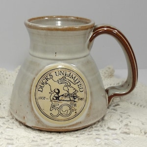 May include: A ceramic mug with a light beige body and a brown handle. The mug features a circular emblem with the text "DUCKS UNLIMITED" and a duck illustration, celebrating its 50th anniversary from 1937 to 1987. The rim and handle are brown.