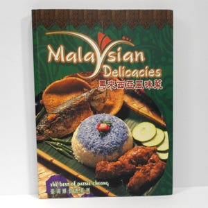 May include: A cookbook titled Malaysian Delicacies featuring a cover image of a blue-tinted rice mound, fried fish, cucumber slices, and chicken on a banana leaf. The text reads Malaysian Delicacies, the best of Patsie Cheong, and Chinese characters.