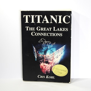 May include: A black hardcover book titled "Titanic: The Great Lakes Connections" by Cris Kohl. The cover features a photo of a ship's bow with a map of the Great Lakes superimposed on it. A gold sticker with the text "Autographed Copy" is on the bottom right corner of the cover.
