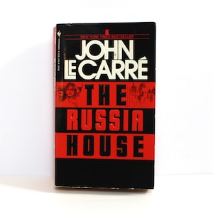 May include: A black and red paperback book with the title "The Russia House" by John le Carre. The book is a New York Times bestseller.