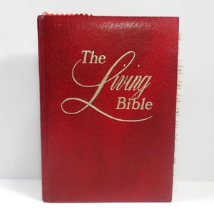 Puede incluir: Un libro rojo de tapa dura titulado "The Living Bible" con letras doradas. El libro tiene una cubierta texturizada y un marcador de página en el lateral. El libro es un texto religioso.