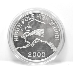 May include: A silver coin encased in a clear plastic holder. The coin features the text "NORTH POLE HIGH SCHOOL" around the top edge and the year "2000" at the bottom. An outline of Alaska and a flag are in the center.