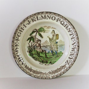 May include: A white ceramic plate with a brown alphabet border and a green and brown illustration of Robinson Crusoe rescuing Friday. The plate has the text "CRUSOE RESCUES FRIDAY" printed on the bottom.