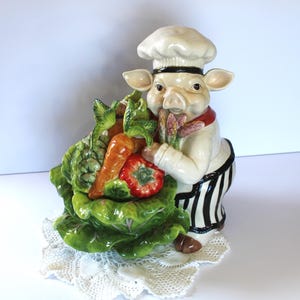 May include: A ceramic pig figurine wearing a chef's hat and holding a basket of vegetables. The pig is wearing a white chef's jacket and black and white striped pants. The vegetables include carrots, lettuce, asparagus, and a tomato.