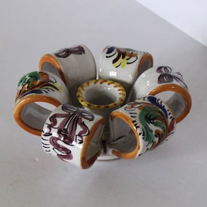 Könnte beinhalten: Ein weißer Kerzenhalter aus Keramik mit sechs Ringen, die jeweils mit bunten Blumenmustern verziert sind. Die Ringe sind in Form einer Blume angeordnet, mit einer zentralen Tasse für die Kerze.