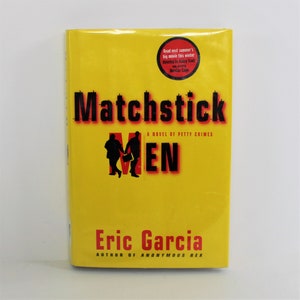 May include: A yellow paperback book with the title "Matchstick Men" in black and red lettering. The book is a novel of petty crimes by Eric Garcia, author of "Anonymous Rex".