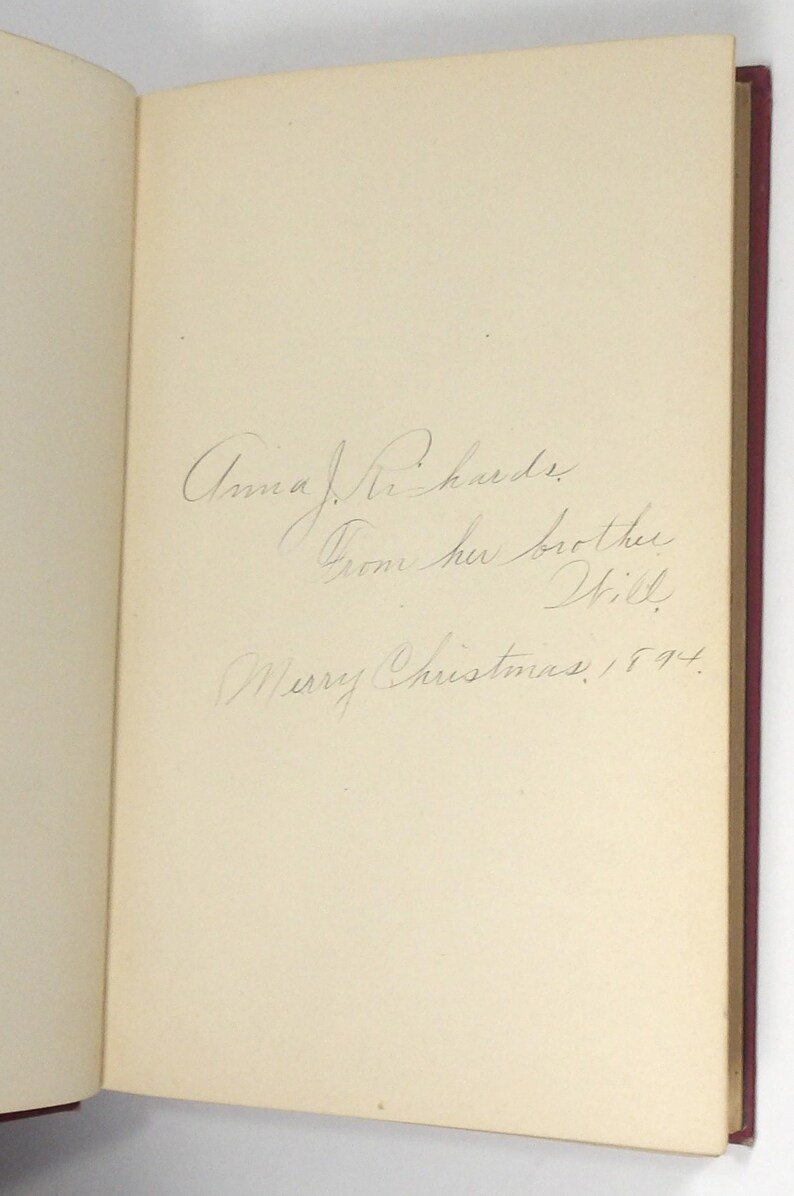 Może przedstawiać: Otwarta książka z odręcznym napisem na prawej stronie. Napis brzmi: "Anna J. Richards, Od jej brata Will, Wesołych Świąt, 1894." Książka ma czerwony grzbiet.