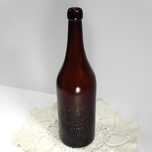 May include: A brown glass bottle with a long neck and a rounded base. The bottle has embossed text and is likely an antique. The bottle is on a white lace doily. The bottle is likely from the early 20th century.