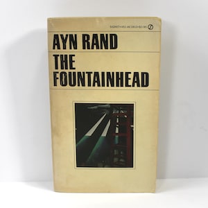 Puede incluir: Portada del libro "The Fountainhead" de Ayn Rand. El libro tiene una cubierta beige con texto negro y una imagen rectangular de un edificio con líneas blancas y acentos verdes y rojos.