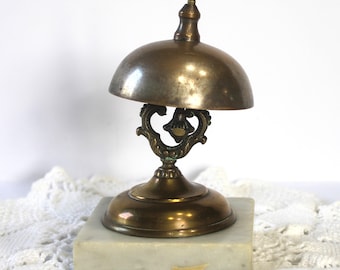 May include: An antique brass bell with a domed top and ornate filigree support, mounted on a square marble base. The bell has a small handle on top. The overall design is vintage and decorative.