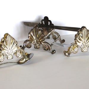 May include: A set of three silver-plated cutlery holders with ornate designs. Each holder features a decorative shell-like pattern and is designed to hold a knife, fork, or spoon.