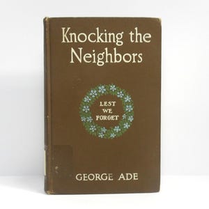 May include: A vintage book titled "Knocking the Neighbors" by George Ade. The cover is brown with white text and a floral wreath design. The text "Lest We Forget" is inside the wreath.
